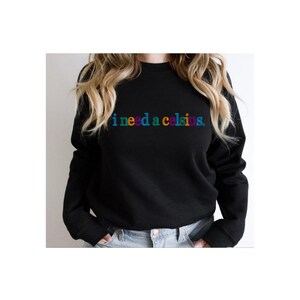 May include: Black crew neck sweatshirt with the phrase "i need a celsius" in rainbow-colored text across the chest. The sweatshirt is a solid black color and has long sleeves.