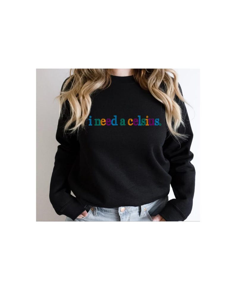 May include: Black crew neck sweatshirt with the phrase "i need a celsius" in rainbow-colored text across the chest. The sweatshirt is a solid black color and has long sleeves.