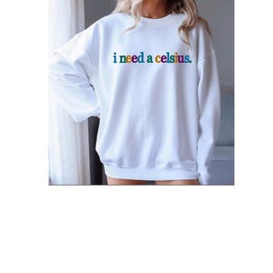 May include: White sweatshirt with the text "i need a celsius" in rainbow colors. The sweatshirt has long sleeves and a crew neck. The text is centered on the chest.