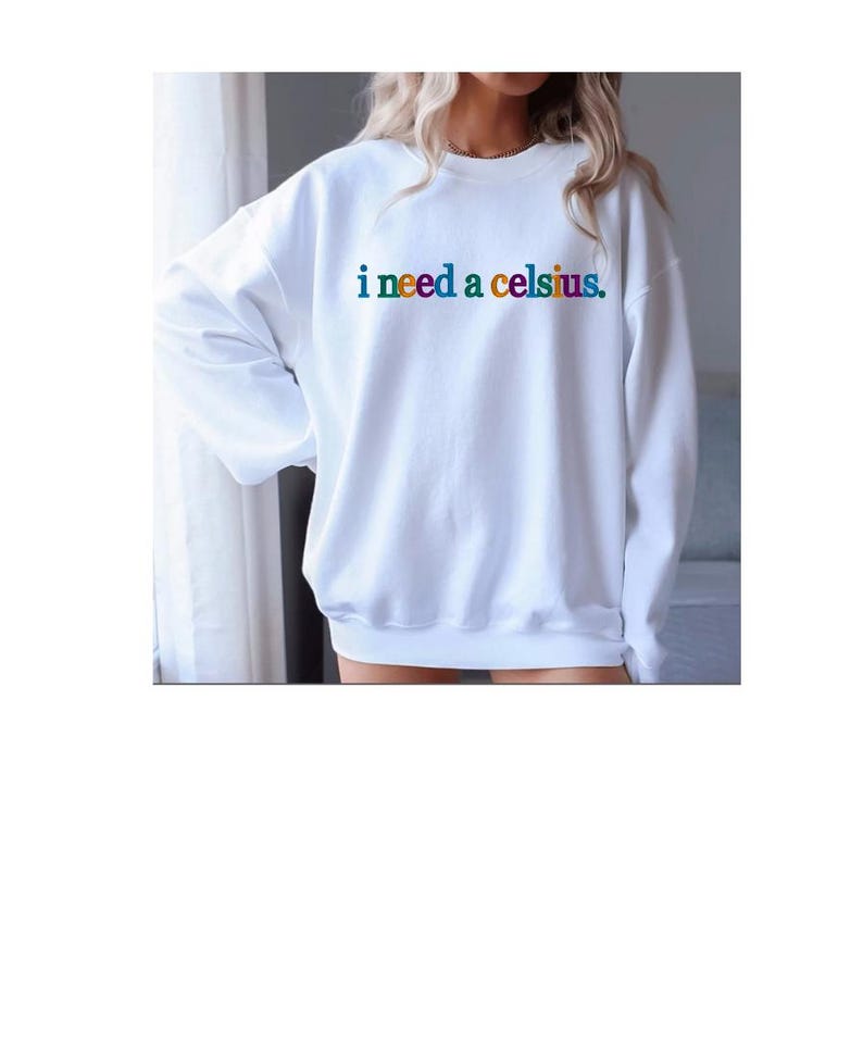 May include: White sweatshirt with the text "i need a celsius" in rainbow colors. The sweatshirt has long sleeves and a crew neck. The text is centered on the chest.