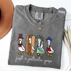 May include: A gray Comfort Colors t-shirt featuring a patriotic goose design. The graphic includes four geese dressed as Uncle Sam, a hot dog, the Statue of Liberty, and a colonial figure. The text "just a patriotic goose" is written in cursive below the geese.
