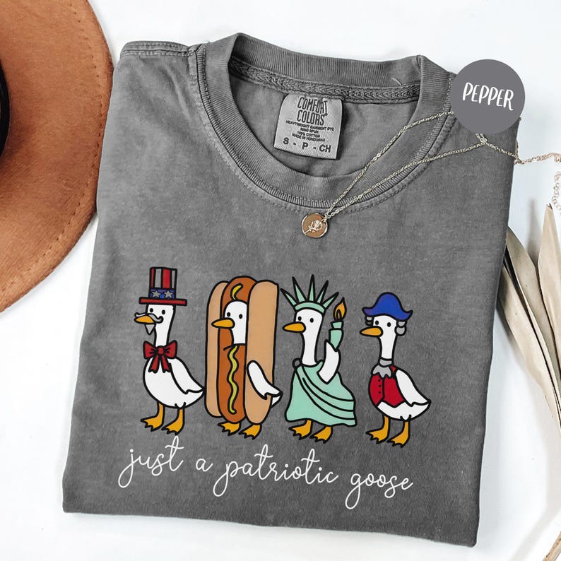 May include: A gray Comfort Colors t-shirt featuring a patriotic goose design. The graphic includes four geese dressed as Uncle Sam, a hot dog, the Statue of Liberty, and a colonial figure. The text "just a patriotic goose" is written in cursive below the geese.