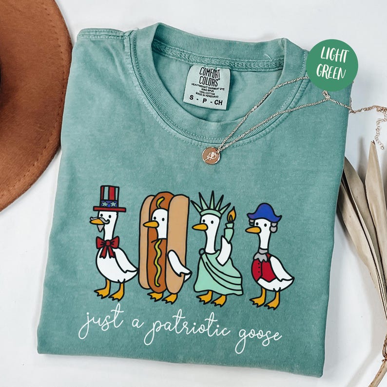 May include: A light green t-shirt featuring a graphic of four cartoon geese dressed in patriotic attire. The geese are depicted as Uncle Sam, a hot dog, the Statue of Liberty, and a colonial figure. The text "just a patriotic goose" is printed below.