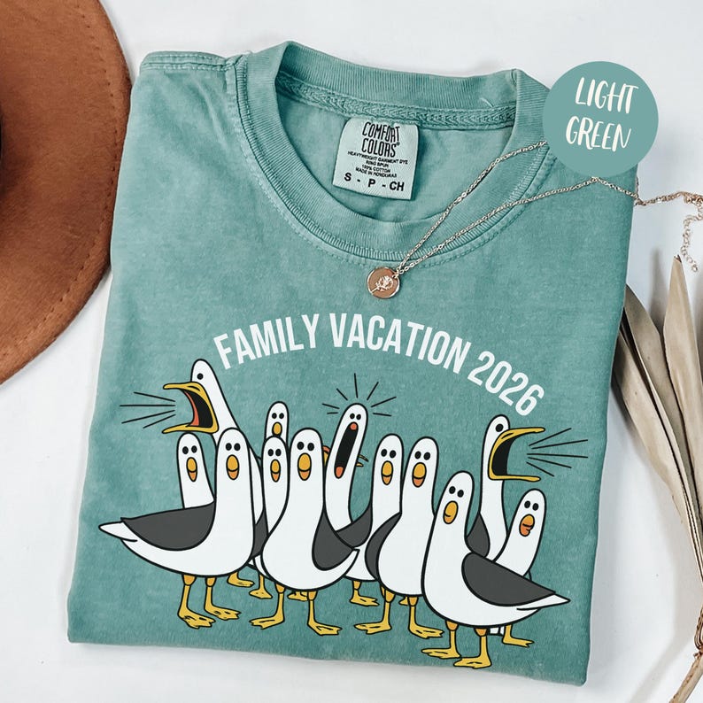 May include: A light green short-sleeve t-shirt featuring a graphic of multiple white seagulls with gray wings and yellow feet. The text "FAMILY VACATION 2026" is printed in white above the birds. The shirt has a crew neckline and a Comfort Colors label.