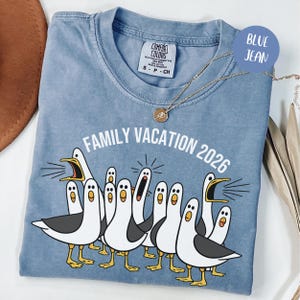 May include: A blue jean colored t-shirt with a crew neckline and a graphic print of multiple white seagulls with grey wings and yellow beaks. The text "FAMILY VACATION 2026" is printed in white above the birds. A gold necklace rests on the shirt.
