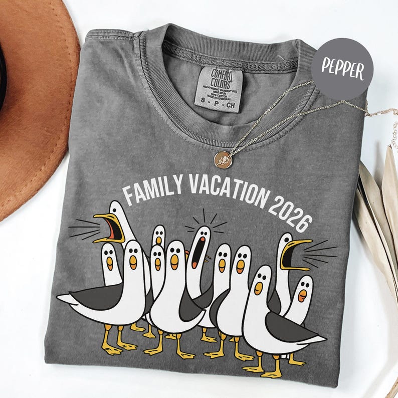 May include: A pepper-colored t-shirt featuring a graphic of multiple white and grey seagulls with yellow beaks and feet. The text "FAMILY VACATION 2026" is printed in white above the birds. The shirt has a Comfort Colors brand label at the neck.