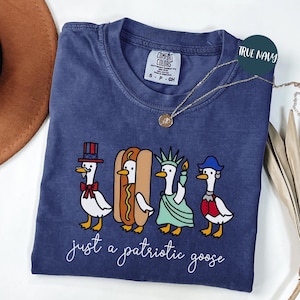 May include: A navy blue t-shirt with a graphic of four cartoon geese dressed in patriotic attire. The geese are wearing a top hat, a hot dog costume, a Statue of Liberty outfit, and a colonial hat. The text "just a patriotic goose" is below the graphic.