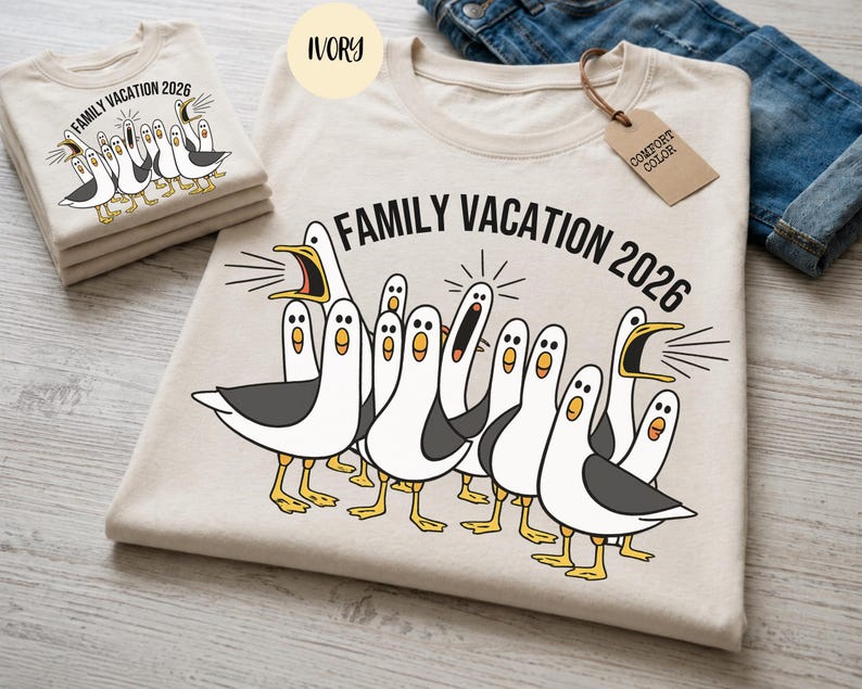 May include: An ivory t-shirt featuring a graphic of multiple white and grey seagulls with yellow beaks and feet. Some seagulls have open beaks as if squawking. The text "FAMILY VACATION 2026" is printed in black above the seagull illustration.