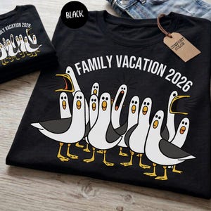 May include: A black t-shirt featuring a graphic of multiple white and gray seagulls with yellow beaks and feet. The text "FAMILY VACATION 2026" is printed in white above the birds. A brown paper tag labeled "COMFORT COLOR" is attached to the collar.