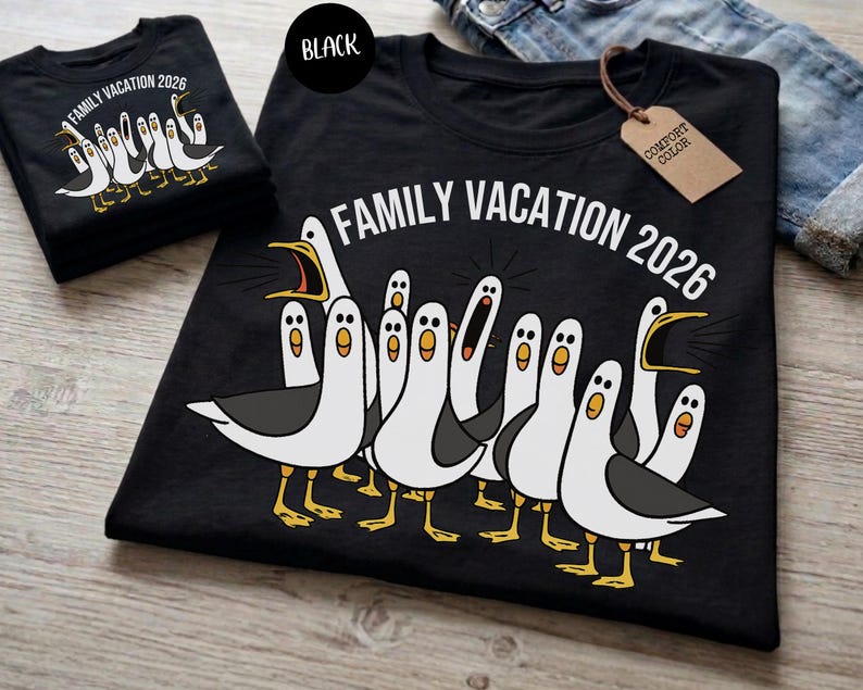 May include: A black t-shirt featuring a graphic of multiple white and gray seagulls with yellow beaks and feet. The text "FAMILY VACATION 2026" is printed in white above the birds. A brown paper tag labeled "COMFORT COLOR" is attached to the collar.