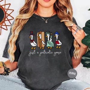 May include: Dark gray t-shirt featuring a whimsical illustration of four geese dressed in patriotic attire: a top hat, a hot dog, the Statue of Liberty, and a red, white, and blue outfit. The text "just a patriotic goose" is written below the geese.