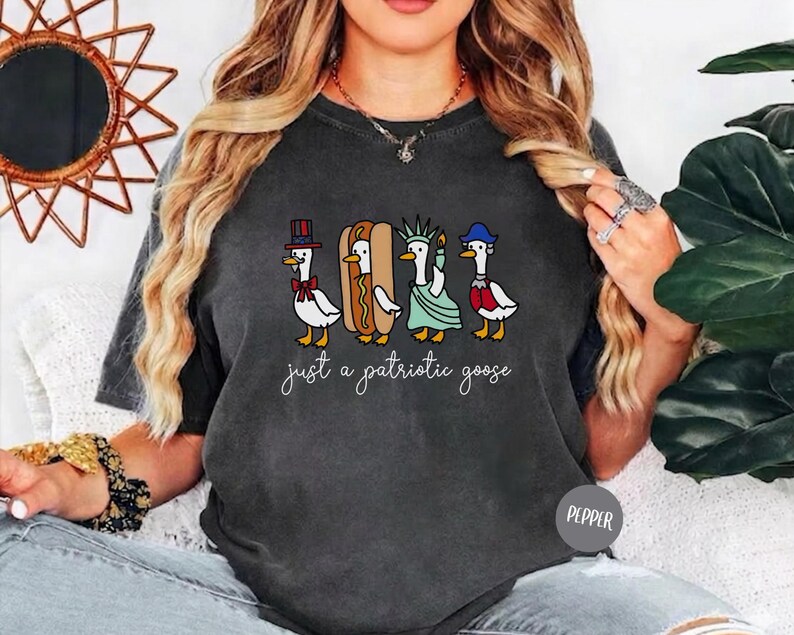 May include: Dark gray t-shirt featuring a whimsical illustration of four geese dressed in patriotic attire: a top hat, a hot dog, the Statue of Liberty, and a red, white, and blue outfit. The text "just a patriotic goose" is written below the geese.