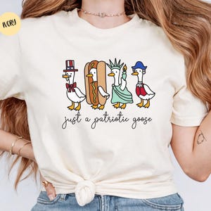 May include: Ivory-colored t-shirt featuring a whimsical illustration of four geese dressed in patriotic attire. One goose wears a top hat, another is a hot dog, a third is the Statue of Liberty, and the last wears a blue hat. The text "just a patriotic goose" is below.