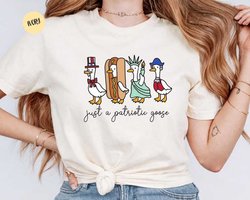 May include: Ivory-colored t-shirt featuring a whimsical illustration of four geese dressed in patriotic attire. One goose wears a top hat, another is a hot dog, a third is the Statue of Liberty, and the last wears a blue hat. The text "just a patriotic goose" is below.