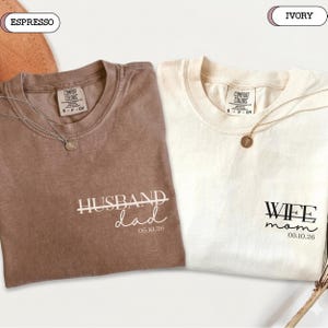 May include: Two Comfort Colors t-shirts, one espresso and one ivory. The espresso shirt says "Dad" with the date "05.10.26". The ivory shirt says "Mom" with the same date. Both shirts have a necklace.