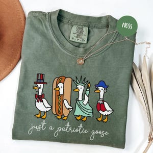 May include: Moss green t-shirt with a patriotic goose design. The graphic features four cartoon geese dressed as patriotic symbols, including a hot dog and the Statue of Liberty. The text "just a patriotic goose" is below the geese.