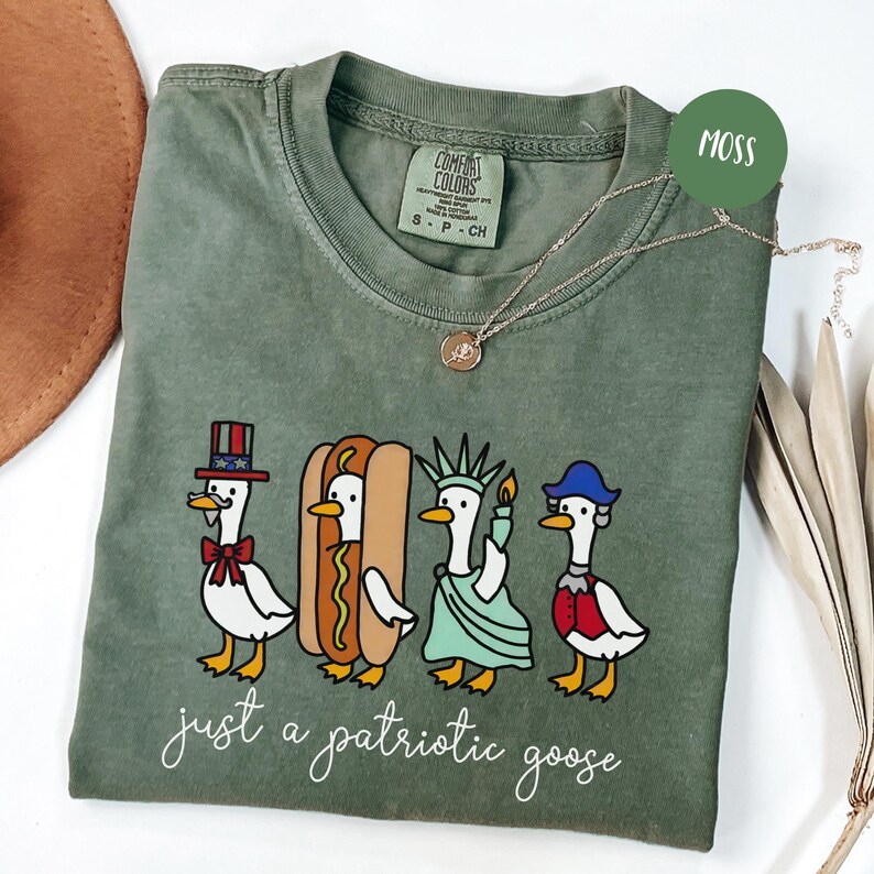 May include: Moss green t-shirt with a patriotic goose design. The graphic features four cartoon geese dressed as patriotic symbols, including a hot dog and the Statue of Liberty. The text "just a patriotic goose" is below the geese.