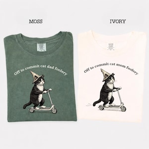 May include: Two t-shirts, one moss green and one ivory, each featuring a graphic of a black and white cat riding a scooter and wearing a wizard hat. The text on the green shirt reads "Off to commit cat dad foolery" and the ivory shirt says "Off to commit cat mom foolery".