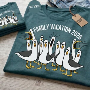 May include: A dark teal t-shirt featuring a graphic of multiple white and gray seagulls with yellow beaks and feet. The text "FAMILY VACATION 2026" is printed in white above the birds. A brown cardboard tag labeled "COMFORT COLOR" is attached.