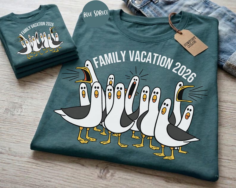 May include: A dark teal t-shirt featuring a graphic of multiple white and gray seagulls with yellow beaks and feet. The text "FAMILY VACATION 2026" is printed in white above the birds. A brown cardboard tag labeled "COMFORT COLOR" is attached.