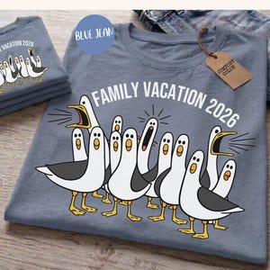 May include: A blue jean colored t-shirt featuring a graphic of multiple white and grey seagulls with yellow beaks and feet. The text "FAMILY VACATION 2026" is printed in white above the birds. A brown tag labeled "COMFORT COLOR" is attached.