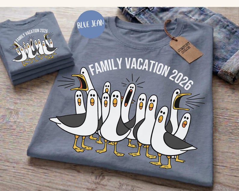 May include: A blue jean colored t-shirt featuring a graphic of multiple white and grey seagulls with yellow beaks and feet. The text "FAMILY VACATION 2026" is printed in white above the birds. A brown tag labeled "COMFORT COLOR" is attached.