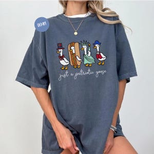 May include: A denim blue t-shirt featuring a whimsical illustration of four geese dressed in patriotic attire. The geese are depicted as a hot dog, the Statue of Liberty, and wearing a top hat and a sailor's hat. The text "just a patriotic goose" is below the geese.