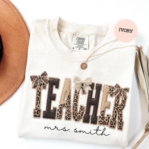 May include: Ivory-colored Comfort Colors t-shirt with the word "TEACHER" in a mix of brown, leopard print, and tan fabric. The name "mrs. smith" is written below the word "TEACHER". A gold necklace and bows are also visible.