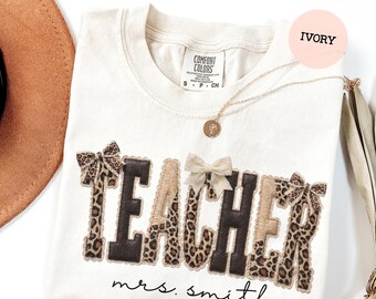 May include: Ivory-colored Comfort Colors t-shirt with the word "TEACHER" in a mix of brown, leopard print, and tan fabric. The name "mrs. smith" is written below the word "TEACHER". A gold necklace and bows are also visible.