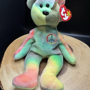 May include: A colorful plush bear with a peace sign on its chest. The bear features a tie-dye design with rainbow hues, including pink, green, yellow, and blue. It has a red tag attached to its ear.