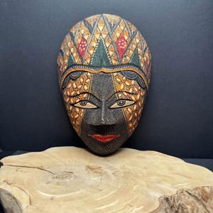 May include: Hand-carved wooden mask with intricate details. The mask features a black face with gold and orange floral patterns, almond-shaped eyes, and red lips. The top of the mask has a geometric design in green, red, and gold. The mask is displayed on a wooden slab.
