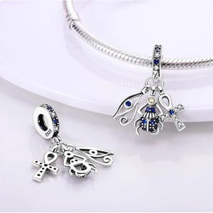 May include: Silver charm bracelet with two charms featuring Egyptian symbols. One charm has a beetle with blue gems, an ankh, and an eye of Horus. The other charm has the same symbols. The charms are detailed and have a vintage look.