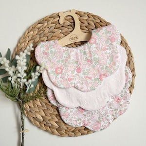 May include: Three baby bibs with scalloped edges and floral patterns in pink and white, displayed on a wooden hanger. The bibs are arranged on a woven, circular placemat, with a sprig of white flowers and greenery to the left.