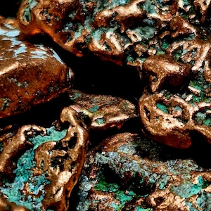 May include: Close-up of several irregularly shaped copper-colored stones with patches of turquoise and green. The stones have a rough, textured surface and are clustered together against a dark background. The image highlights the natural colors and textures of the stones.