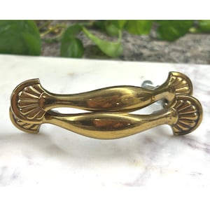 May include: A pair of gold-toned metal drawer pulls featuring a curved, elongated center and fan-shaped ends with embossed radial lines. The hardware has a polished finish and includes a visible metal mounting screw attached to the back side.