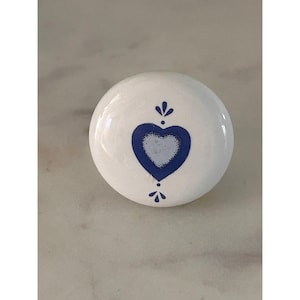 May include: A round white ceramic cabinet knob featuring a central blue heart motif. The heart design includes a lighter blue center and is accented by small blue decorative flourishes positioned directly above and below the heart shape.
