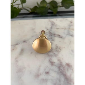 May include: A round cabinet knob with a smooth, brushed gold finish. The knob features a simple, circular face that curves outward, connected to a short, cylindrical metal stem and a small circular base plate for mounting to a drawer or door.
