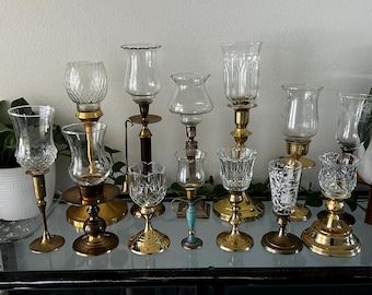 May include: A collection of vintage candle holders in various styles. The holders feature clear glass shades and ornate brass or gold-toned bases. Some have intricate cut glass designs, while others have simple, elegant shapes.
