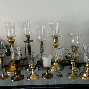 Puede incluir: Una colección de candelabros antiguos con pantallas de vidrio transparente y bases decorativas. Las bases son de oro, latón y madera oscura. Algunos tienen detalles de vidrio grabado, otros diseños texturizados o estampados. Los candelabros están sobre una superficie de vidrio.