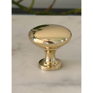 May include: A polished gold-tone cabinet knob featuring a smooth, rounded mushroom-shaped head and a tapered cylindrical base. The metallic surface has a high-gloss finish that reflects light, creating bright highlights across the curved top.