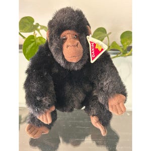 May include: A plush gorilla toy with black synthetic fur and a tan fabric face, hands, and feet. The stuffed animal features a triangular white tag with a red border and the text DAKIN printed in white, attached to the right side of the head.