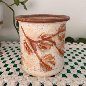 Pode incluir: Um vaso cilíndrico de cerâmica com uma base esbranquiçada salpicada e uma borda marrom. O vaso apresenta um desenho pintado à mão de folhas e galhos marrons. O vaso tem aproximadamente 10 cm de altura e 8 cm de diâmetro.