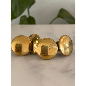 May include: A set of four circular, polished brass buttons with a smooth, reflective surface. Each button features a domed top and a simple, rounded design, showcasing a metallic gold finish that catches the light with visible reflections.