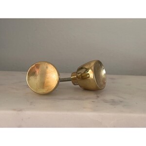 May include: A polished gold-toned metal cabinet knob featuring a smooth, rounded, concave front face and a tapered cylindrical base with a threaded metal screw extending from the back for installation purposes on furniture or cabinetry doors.