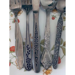 May include: An assortment of vintage silverware, including forks and spoons, arranged on a floral patterned plate. The silverware features various decorative designs, including floral and leaf patterns. The pieces are made of a shiny, silver-colored metal.
