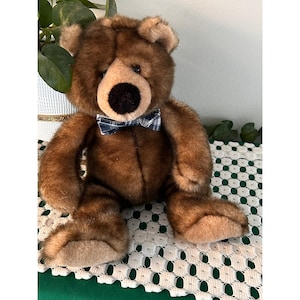 May include: A brown teddy bear with a black nose and a blue and white plaid bow tie. The plush toy has a soft, furry texture and is sitting on a white crocheted surface.