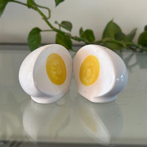 May include: Two white ceramic salt and pepper shakers shaped like halved hard-boiled eggs. Each shaker has a bright yellow yolk design in the center. The shakers are on a reflective surface with a green plant in the background.