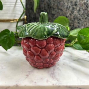 May include: A ceramic lidded jar shaped like a raspberry, featuring a textured, glossy red fruit body and a smooth, dark green leaf-shaped lid with a central stem. The piece has a rounded, bulbous form with realistic, bumpy surface detailing.