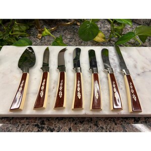 May include: A set of seven cheese knives with stainless steel blades and handles featuring dark wood grain inlays with a light-colored leaf pattern. The collection includes various blade shapes, including a spade, a pointed tip, and a serrated edge.