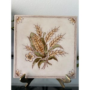 May include: A square ceramic tile featuring a central illustration of a corn cob surrounded by wheat stalks and small flowers in shades of brown, green, and cream. The tile has a thin brown border and a small floral motif in each of the four corners.